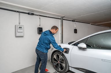 Homme branchant une voiture électrique blanche sur une borne de recharge murale, à côté du compteur électrique, dans un garage