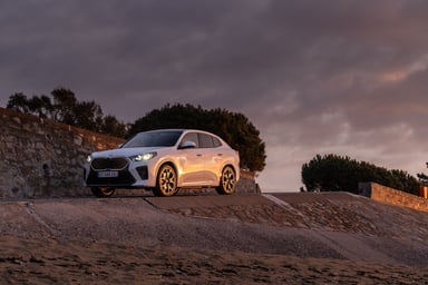 BMW iX2 blanche phares allumés au coucher du soleil sur une promenade côtière