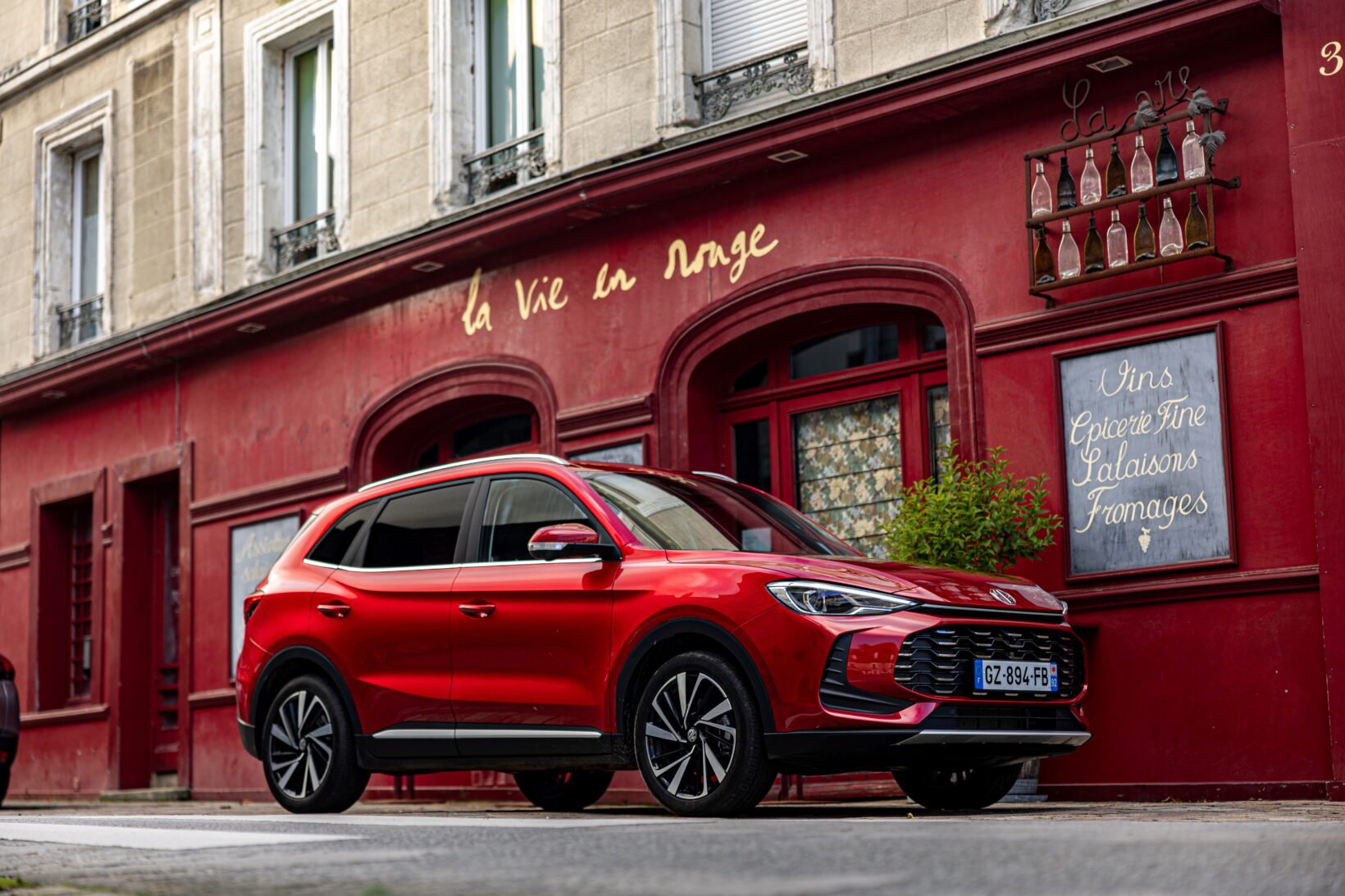 MG ZS Hybrid+ 2026 rouge de trois quarts avant, garé devant une façade de commerce rouge