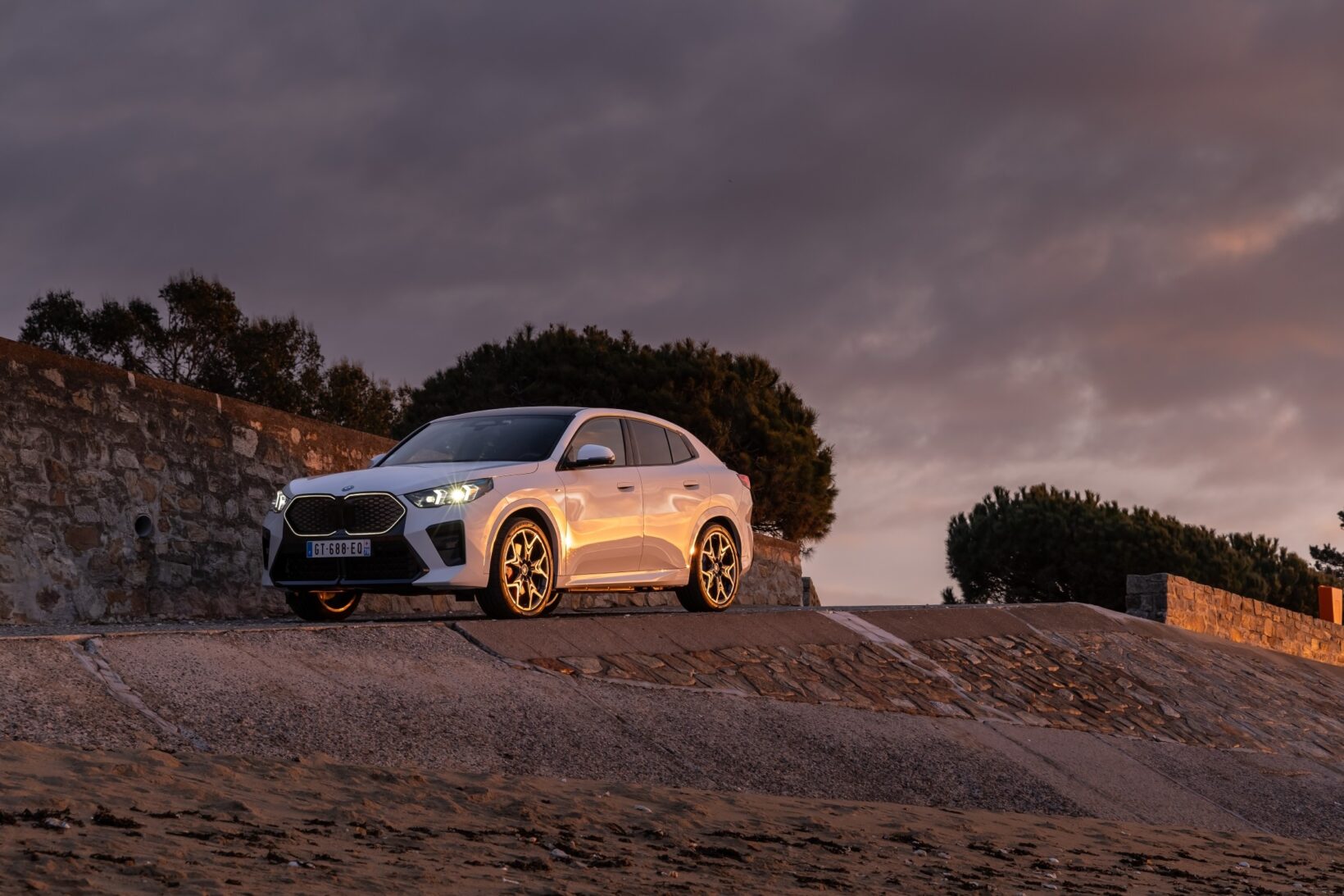 BMW iX2 blanche phares allumés au coucher du soleil sur une promenade côtière