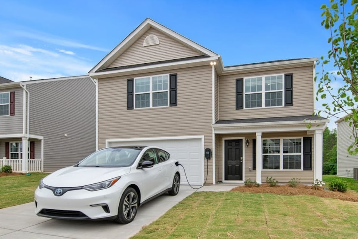 Borne de recharge murale de type wallbox installée sur la façade d'une maison et branchée à une voiture électrique blanche.
