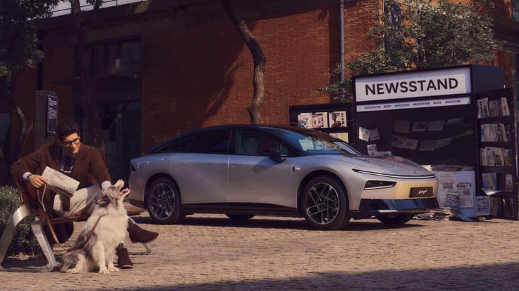 XPeng P7+ grise garée en ville près d’un kiosque à journaux, avec un homme assis et son chien