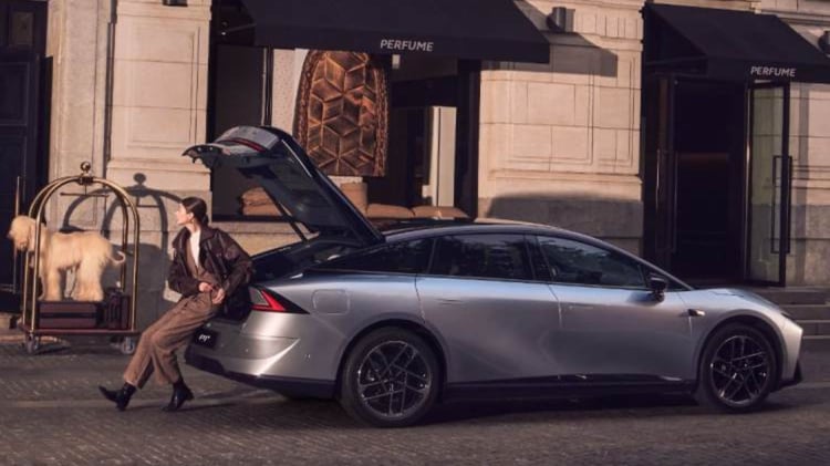 XPeng P7+ grise de profil avec hayon ouvert devant un hôtel, à côté d’un chariot à bagages et d’un chien